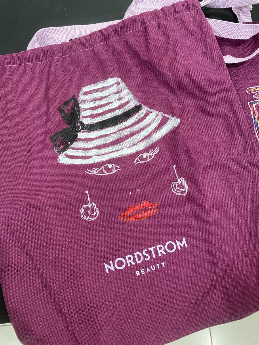 Hand-painted fashion illustration featuring a striped hat, red lips, and earrings on a purple Nordstrom Beauty tote bag for a NYC brand activation.