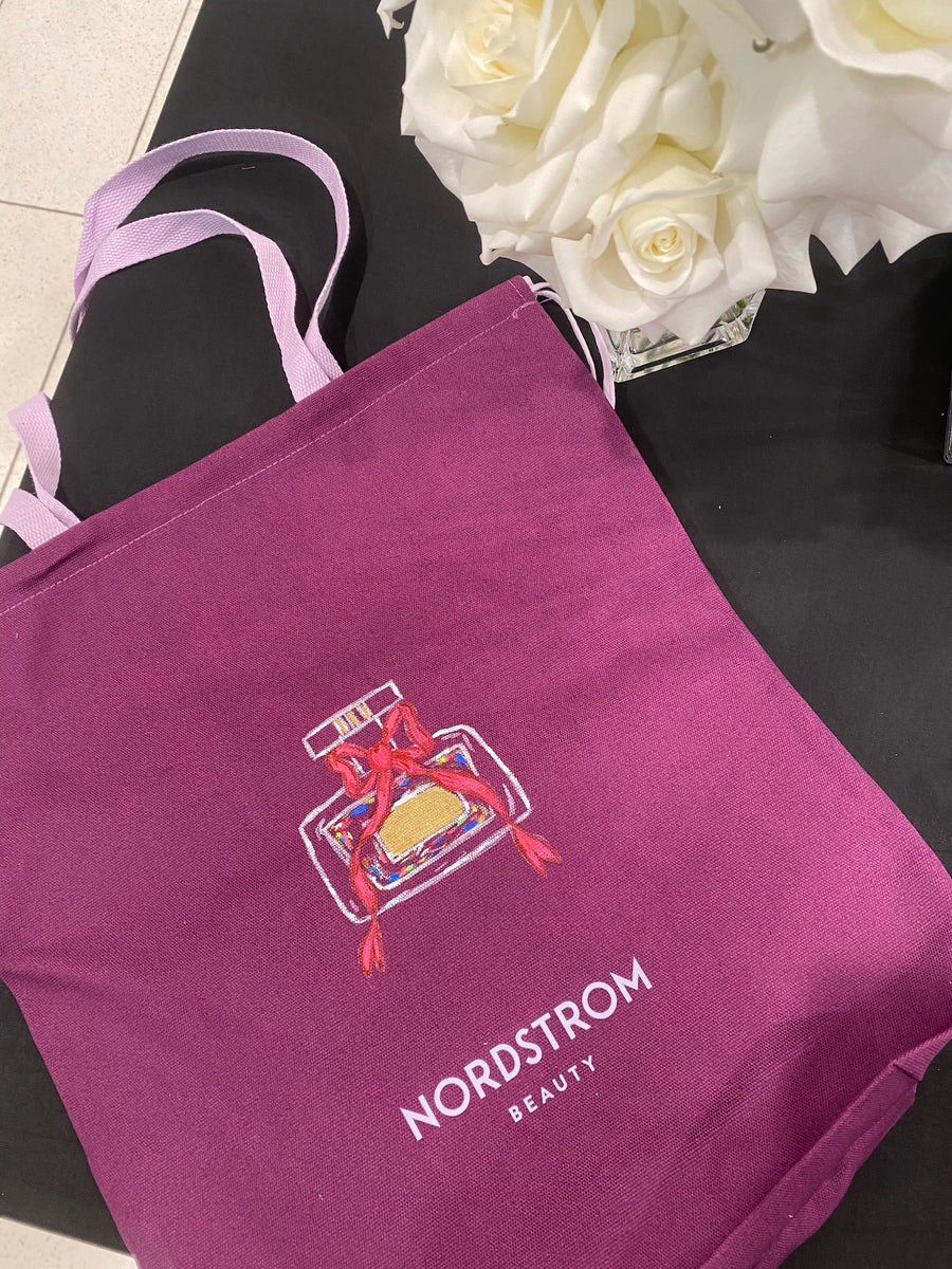 Purple Nordstrom Beauty canvas tote bag with a custom hand-painted perfume bottle illustration displayed beside white roses at a New York City event.