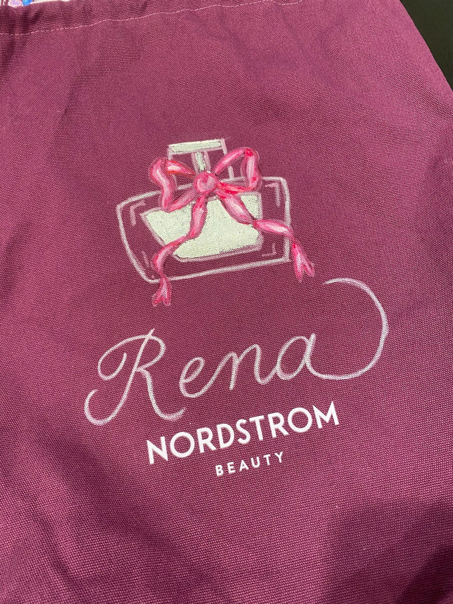 White calligraphy spelling Rena beneath a hand-painted perfume bottle with a pink bow on a personalized Nordstrom Beauty tote bag.