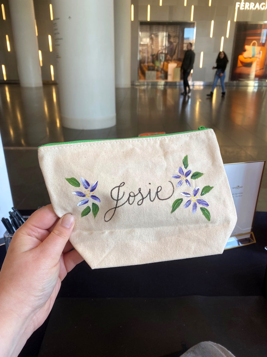 Canvas pouch personalized with the name Josie in calligraphy and purple hand-painted flowers at a Brookfield Place live event.