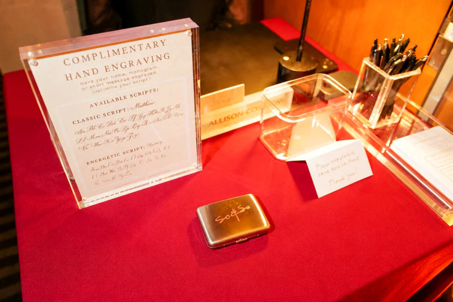 Elegant live engraving station with personalized cigarette cases, lettering samples, and signage at So & So's Piano Bar in NYC.
