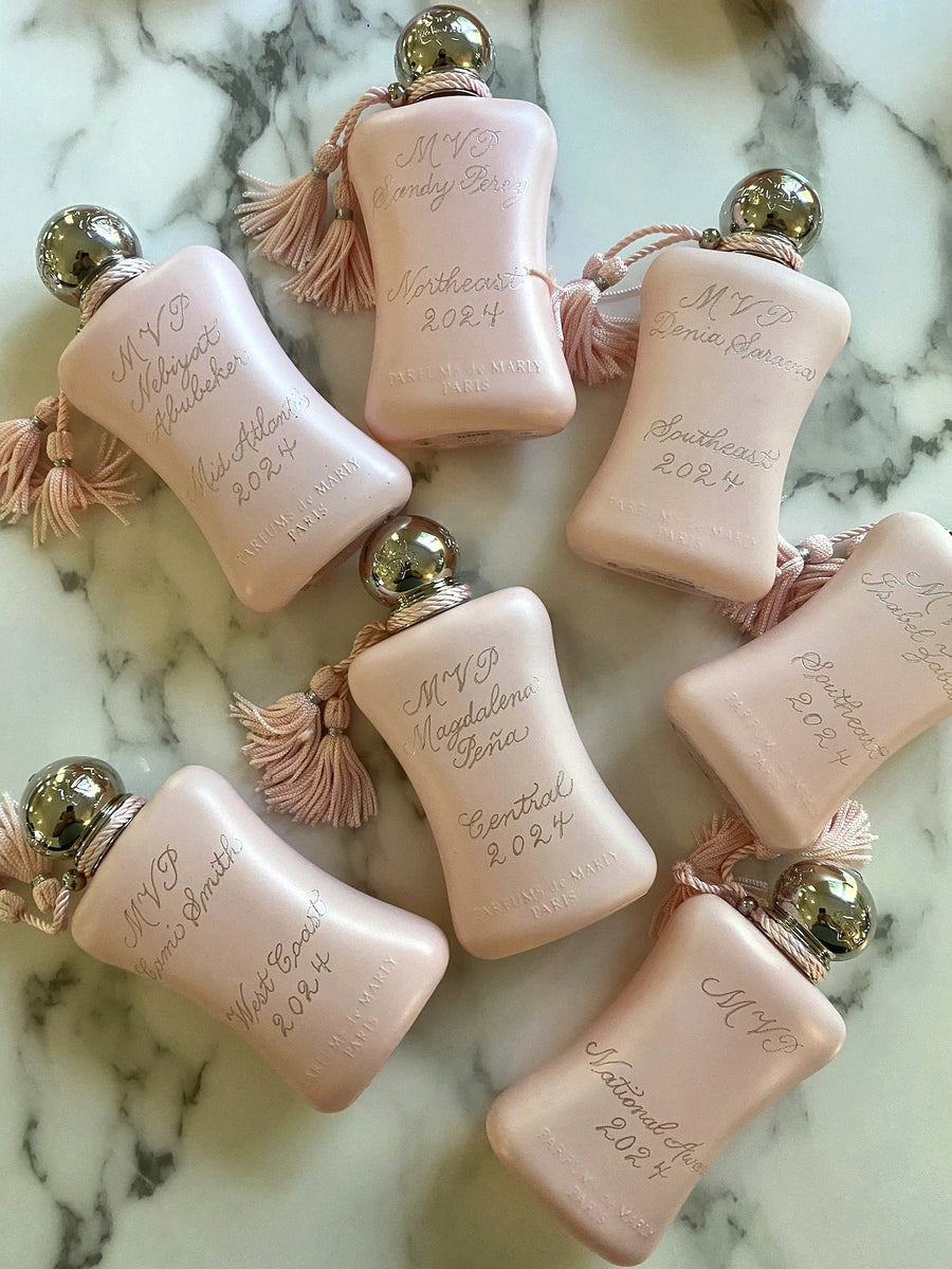 Six pink Parfums de Marly Delina fragrance bottles with hand engraved calligraphy, award titles and dates arranged on a marble surface.